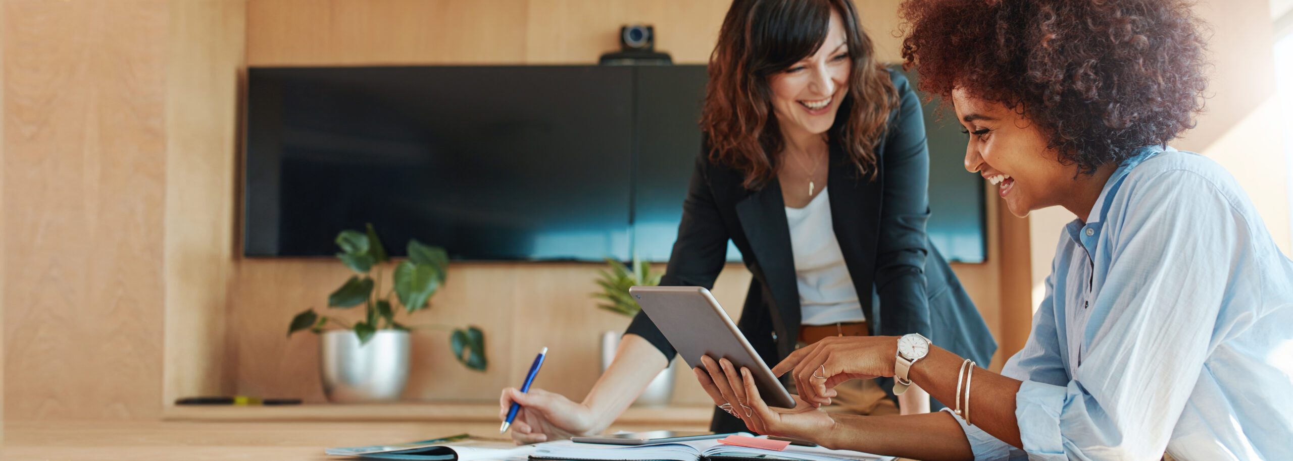 Shot,Of,Two,Businesswoman,Working,Together,On,Digital,Tablet.,Creative Two women in an office setting smiling and discussing something on a tablet. One is sitting, and the other is standing, holding a pen. A television and potted plant in the background.